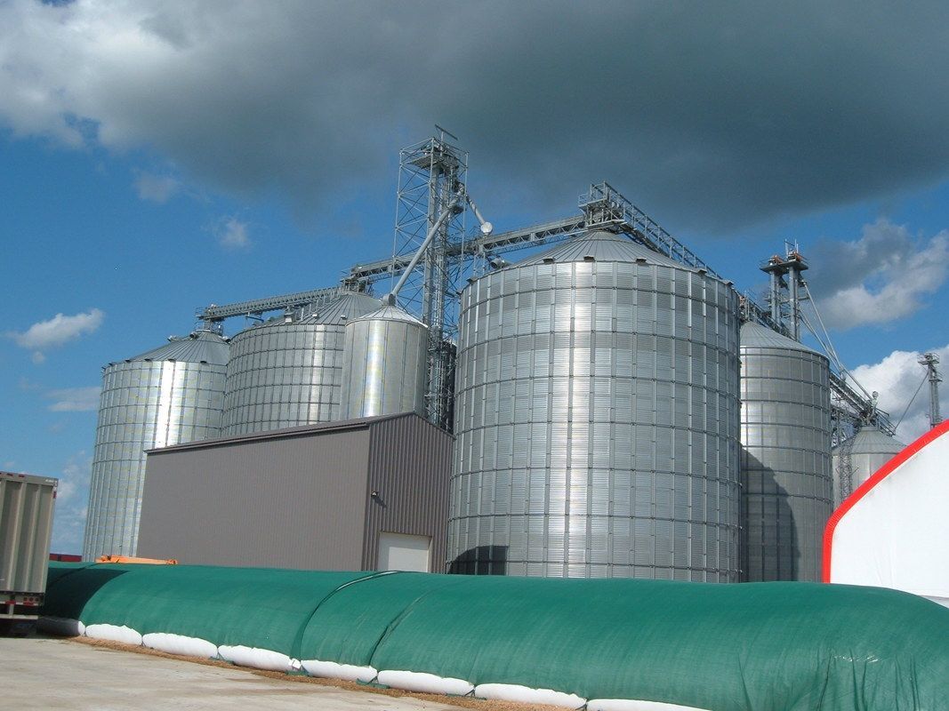 Large metal grain silos at an industrial facility under a cloudy sky