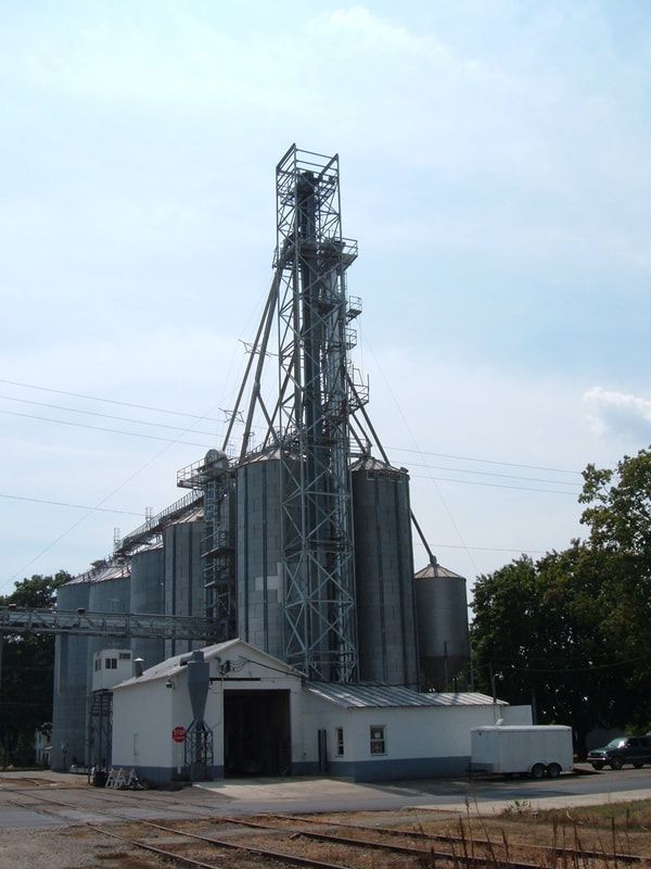 Industrial grain elevator facility with tall metal silos and conveyor tower under a blue sky