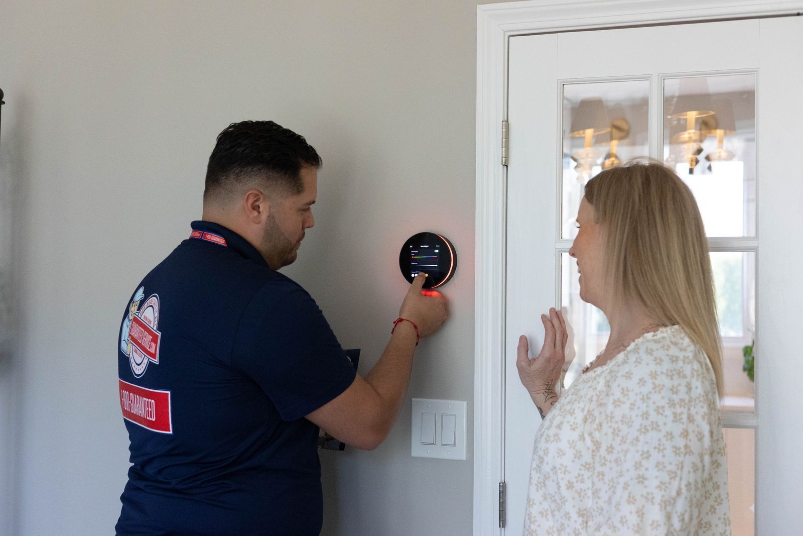 Technician installing a smart thermostat while a woman watches beside a white door.