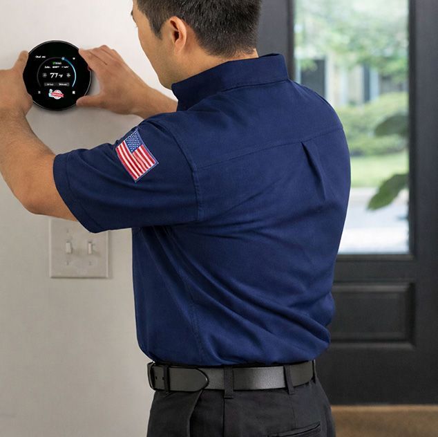 Person adjusting a round thermostat on a wall beside a door, wearing a navy shirt with an American flag patch.