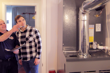 Two people in a doorway beside a large furnace and ductwork in a utility room