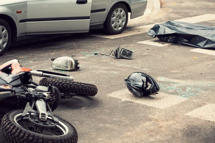 Wrecked motorcycle, helmet, and debris on ground with silver car in background. 