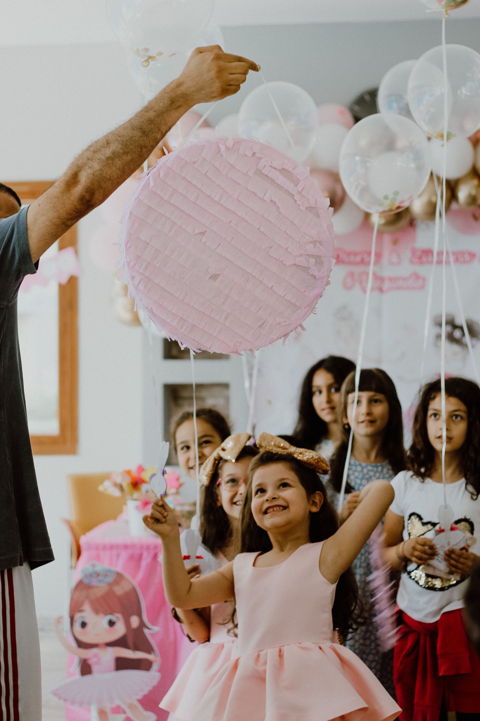 Niños intentando alcanzar un farolillo de papel rosa en una fiesta, con globos y adornos al fondo