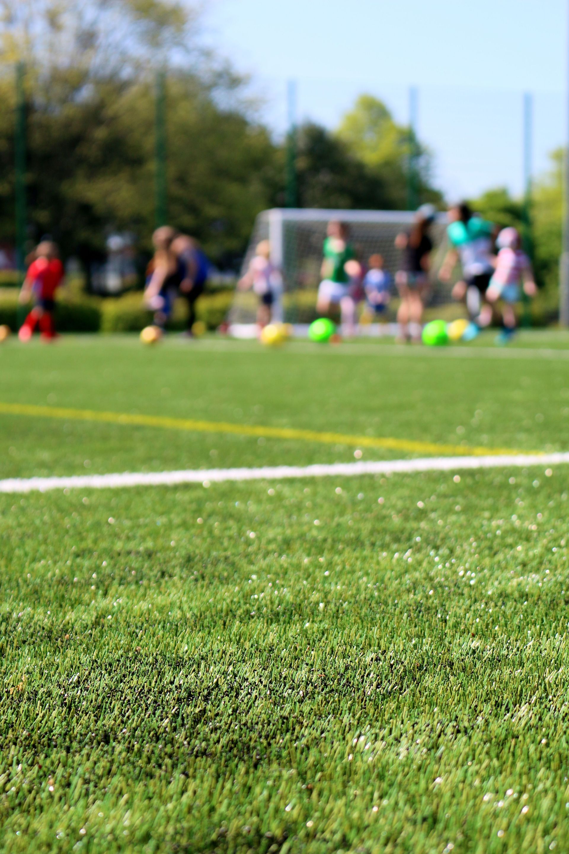 Futbolistas entrenando en un campo cerca de una portería, con césped verde en primer plano y árboles al fondo.