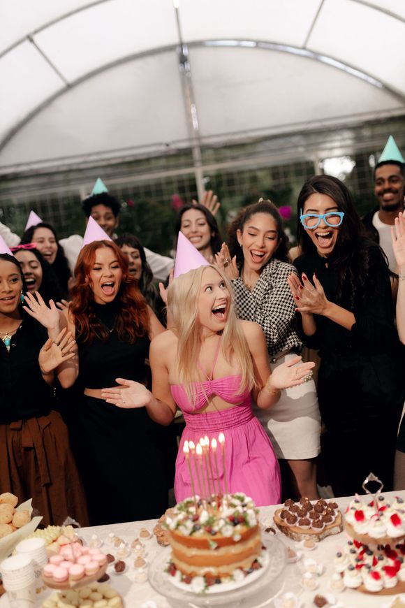 Unos amigos celebran alrededor de una tarta de cumpleaños mientras una mujer con un vestido rosa festeja en una mesa de fiesta