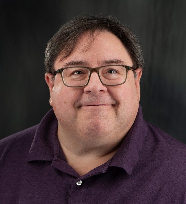 Smiling person wearing glasses and a purple shirt against a dark studio background
