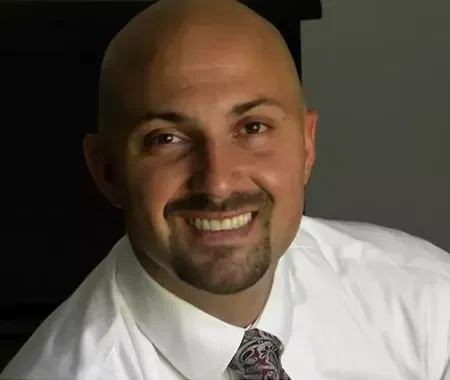 A bald man with a beard is wearing a white shirt and tie and smiling.