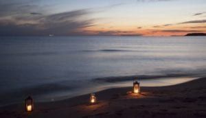 Lanterns glow on a dark beach at dusk, with calm water and a colorful sunset on the horizon