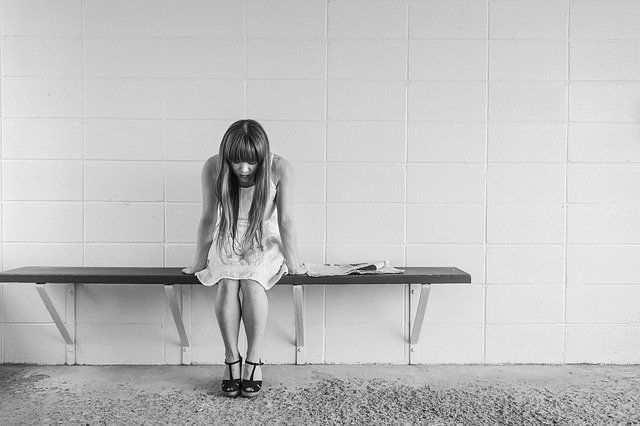 A girl sits hunched on a bench against a tiled wall, looking down in a black-and-white scene.