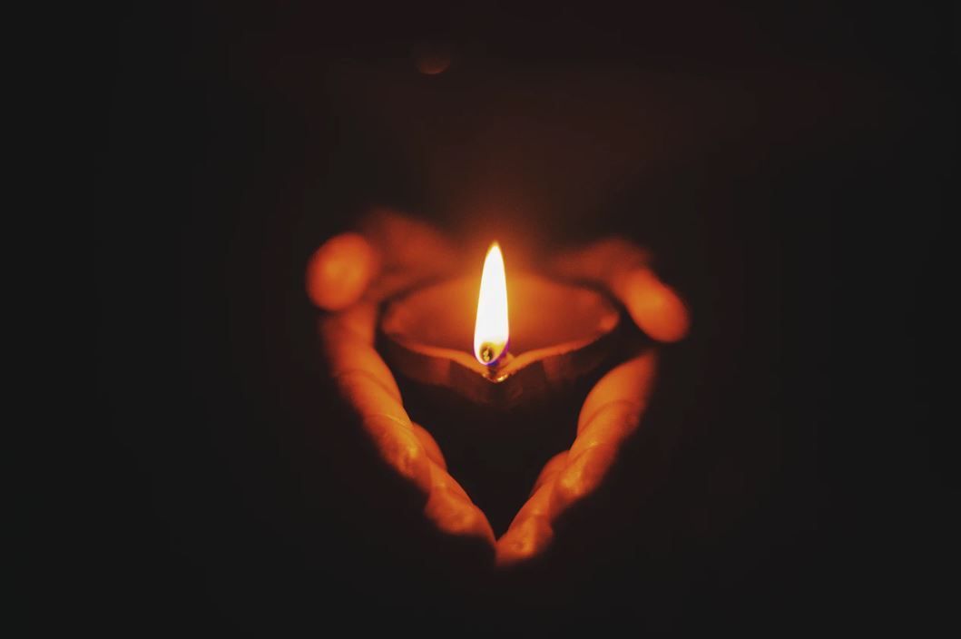 Hands holding a lit candle shaped like a heart in a dark setting