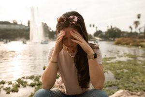 Person sitting by a lake with hands on their face, looking upset near a fountain at sunset