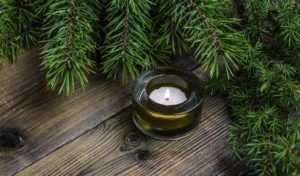 Small white candle in a glass holder on weathered wooden boards beneath evergreen branches.