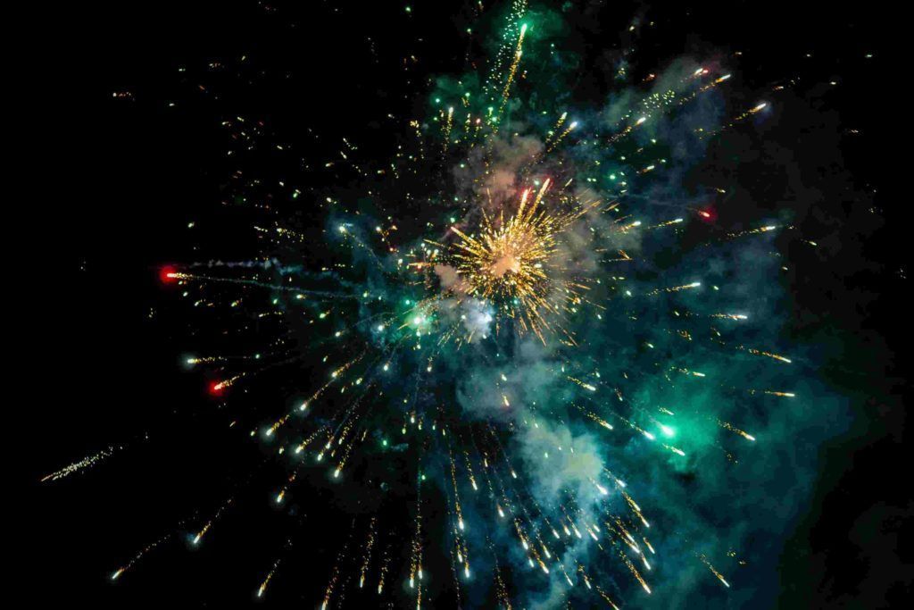 Colorful fireworks burst against a black night sky, with teal, gold, and red sparks radiating outward.