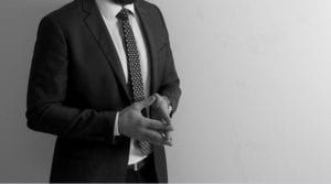 Man in a dark suit and tie standing with hands clasped against a plain light background
