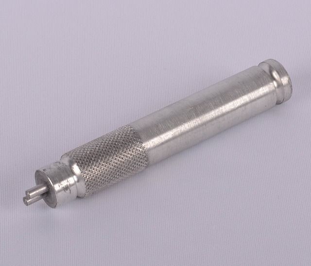 Metal cylindrical tool with knurled grip and small protruding pin, on a light gray background