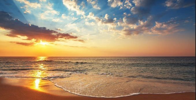 Sunset over a calm ocean with golden light, gentle waves, and a partly cloudy sky