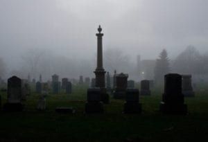 Foggy cemetery with dark gravestones and a tall monument in the center.