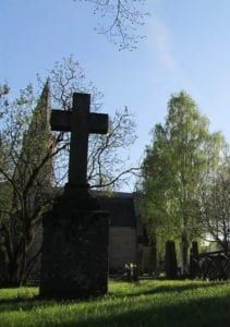 Large stone cross monument in a grassy cemetery under a blue sky, with trees and a small building nearby