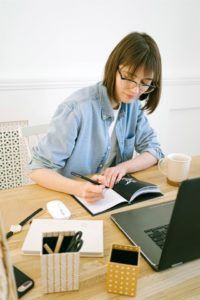 Person writing in a notebook at a desk with a laptop, mug, and stationery.