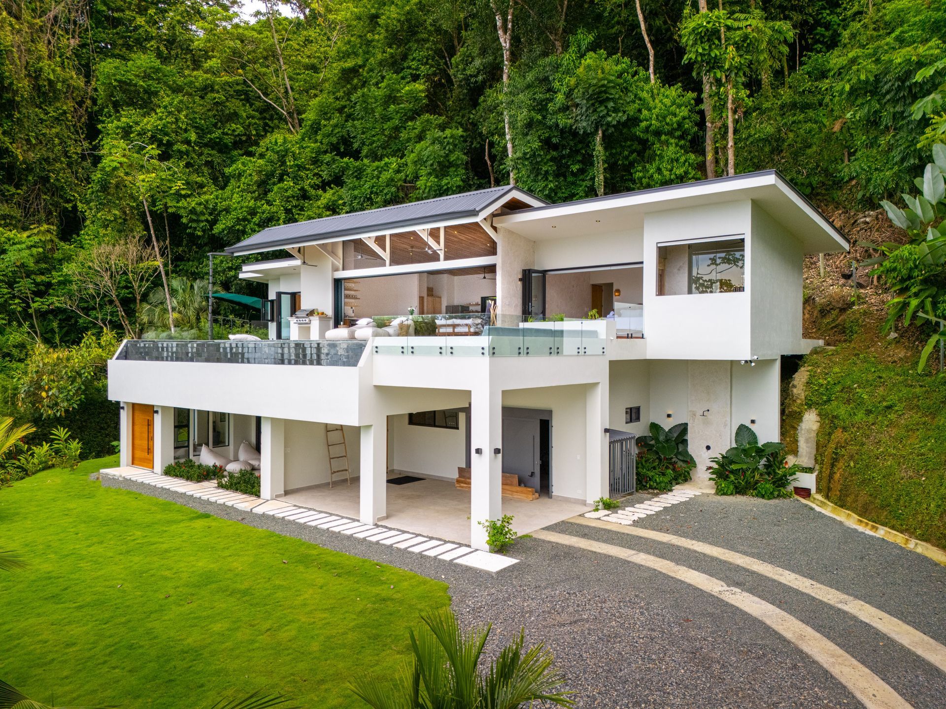 Modern white hillside villa with terrace, green lawn, and lush forest backdrop