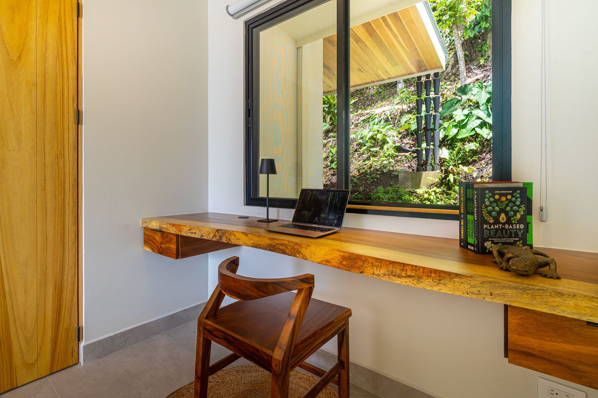 Minimalist desk by a window with a wooden stool, a small laptop, and greenery outside