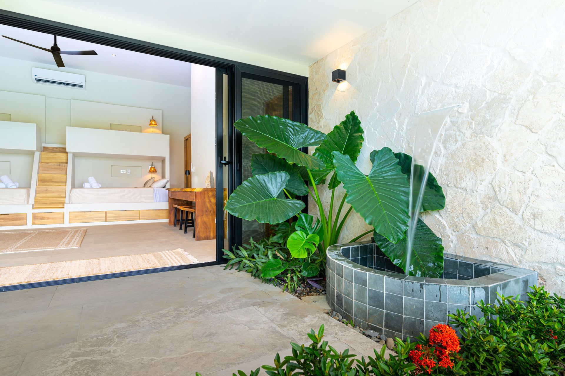 Covered patio with tropical plants, a stone planter, and a view into a bright modern interior room