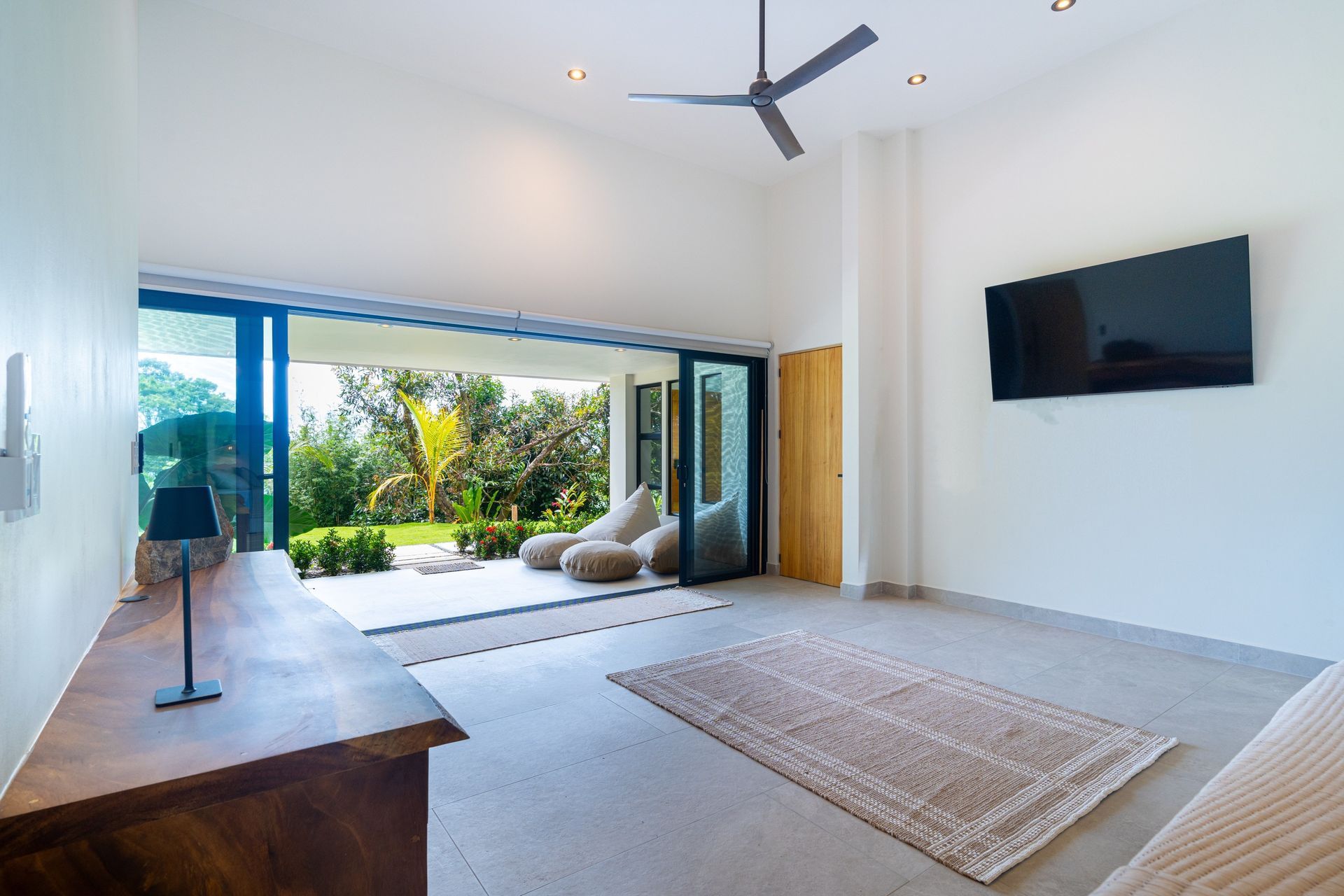 Bright modern living room with sliding glass doors, rug, ceiling fan, and wall-mounted TV, opening to a patio.
