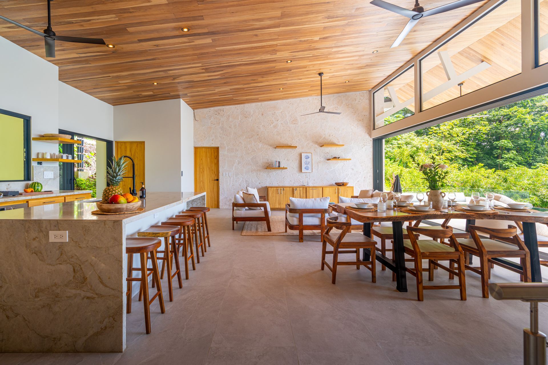Open-air restaurant with wooden tables and stools, stone bar, and greenery beyond the patio opening