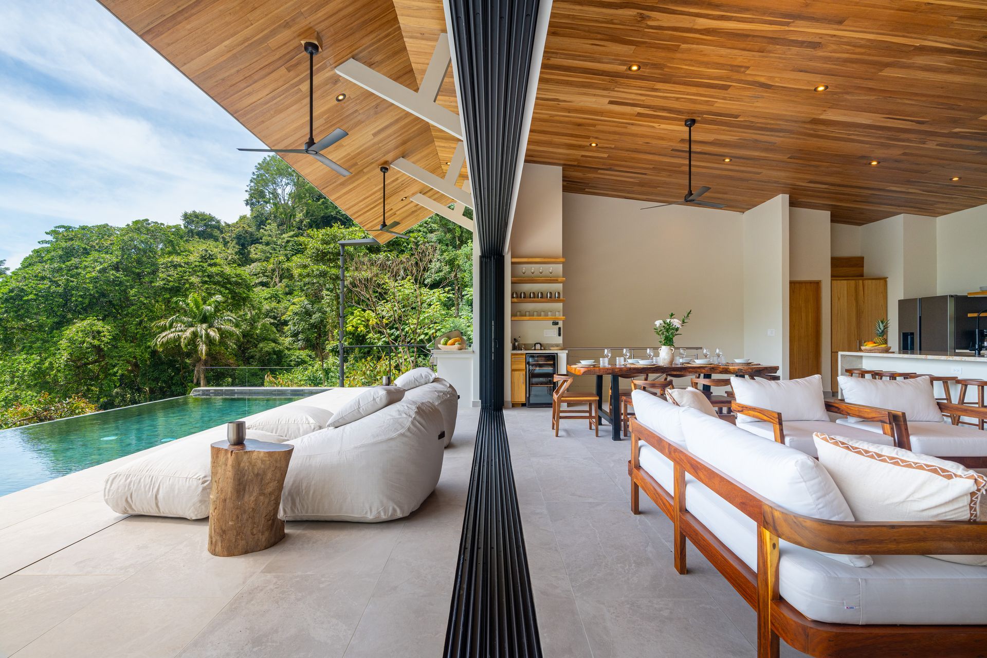 Open-air living room with pool and tropical trees, featuring wood furniture and a high wooden ceiling