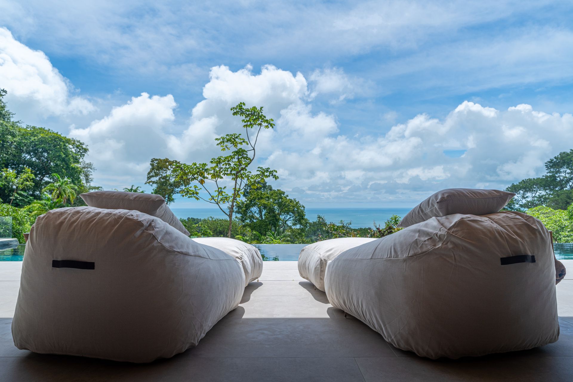 Two white lounge chairs facing a tropical ocean and blue sky from a shaded patio