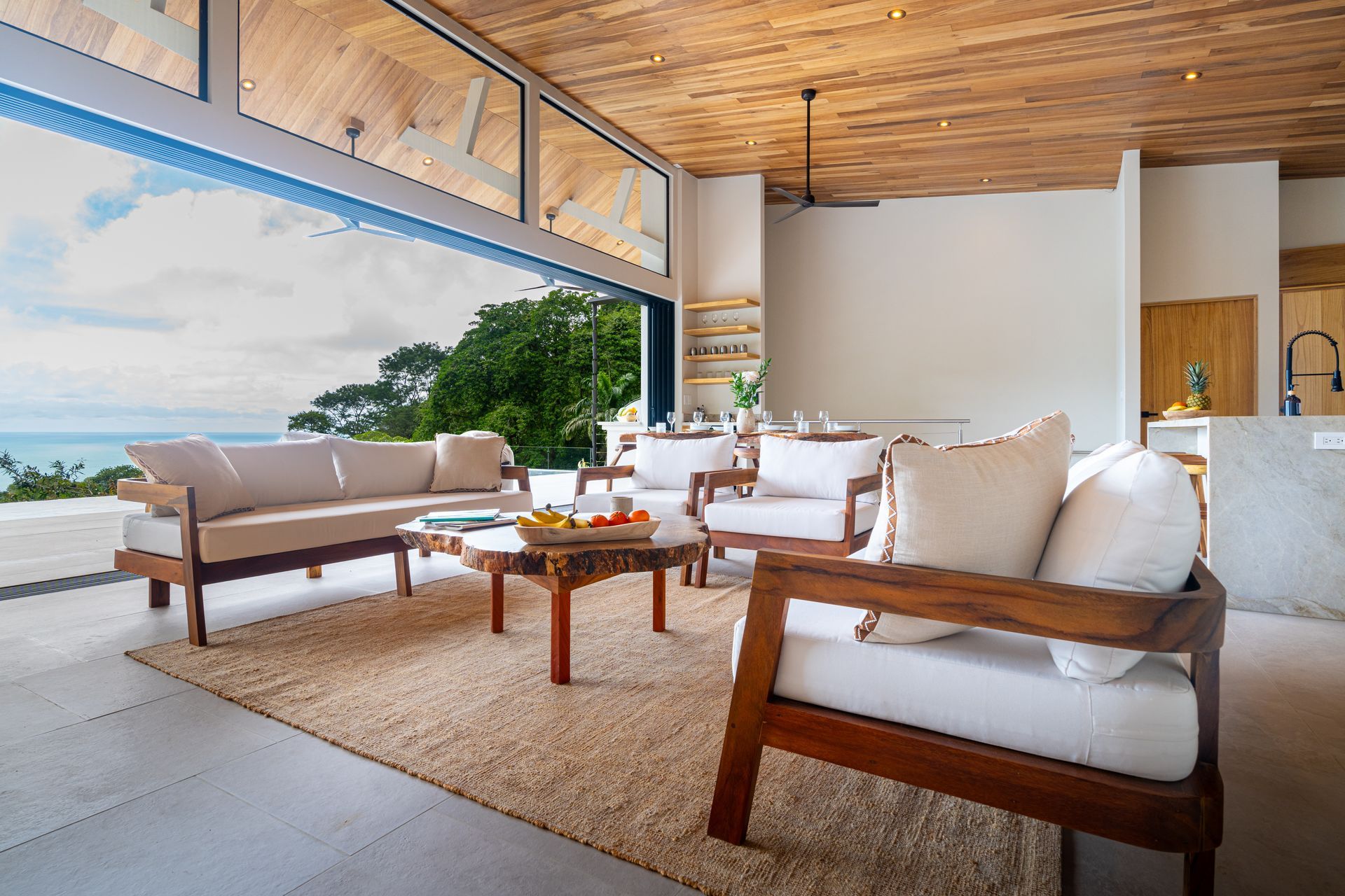 Modern living room with open sea view, white sofas, wooden chairs, and woven rug under a wood ceiling
