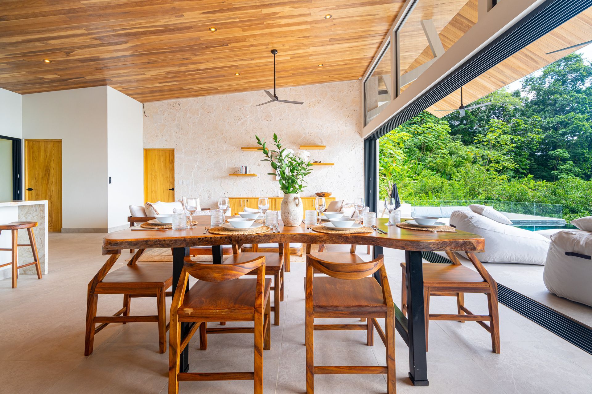 Open-air patio dining area with wooden table and chairs, sofa, and green garden view
