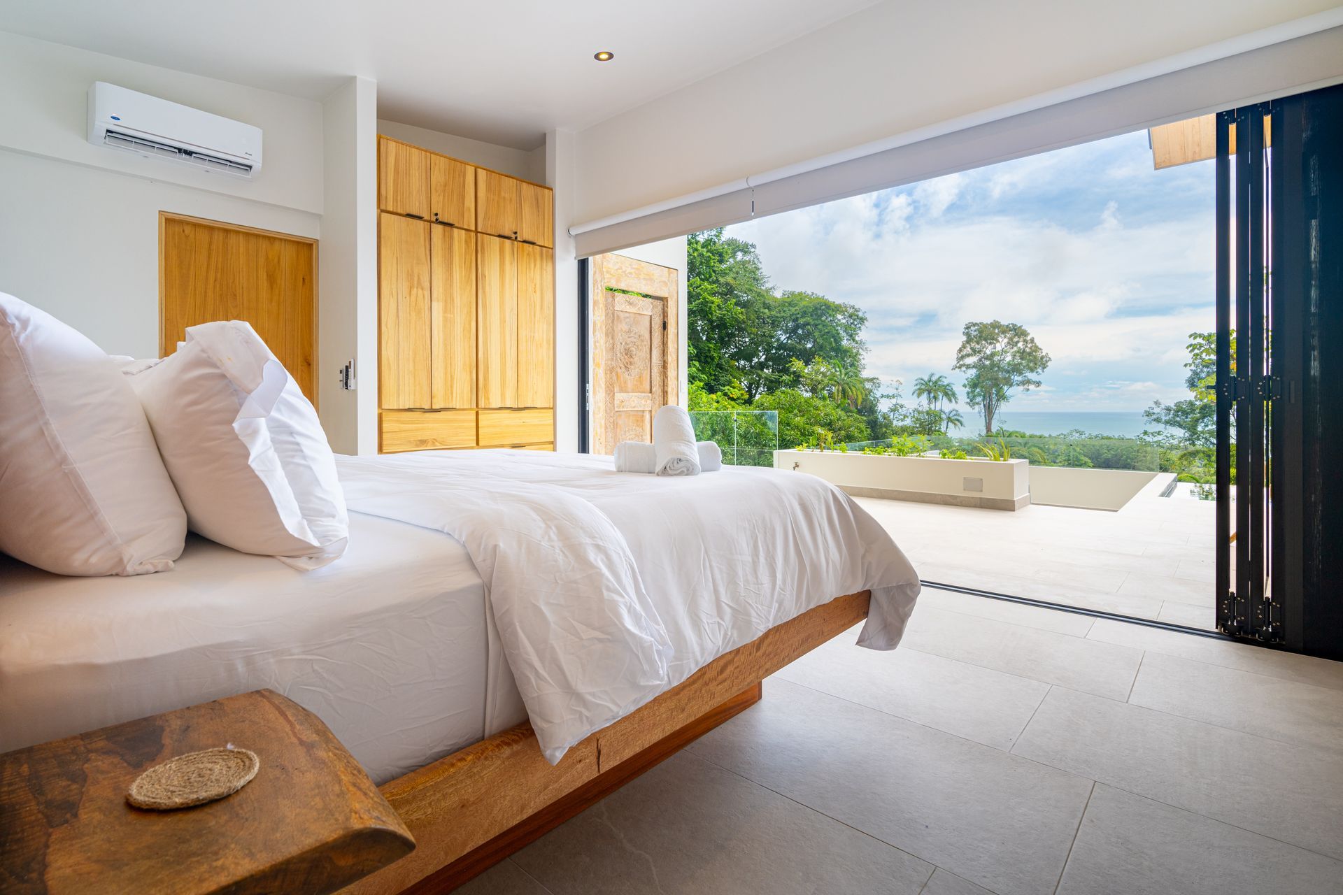Bright bedroom with a wooden bed, open sliding doors, and a view of green hills and ocean beyond.