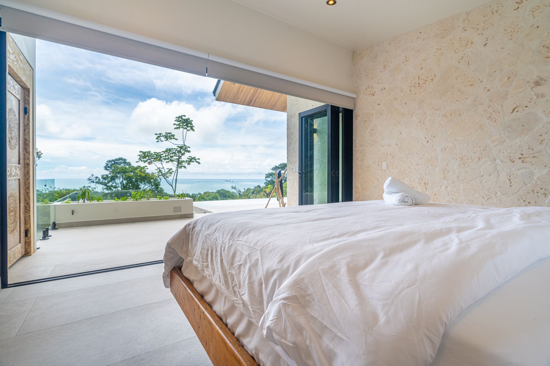 Bright bedroom with white bed, open sliding doors, and ocean view beyond a balcony