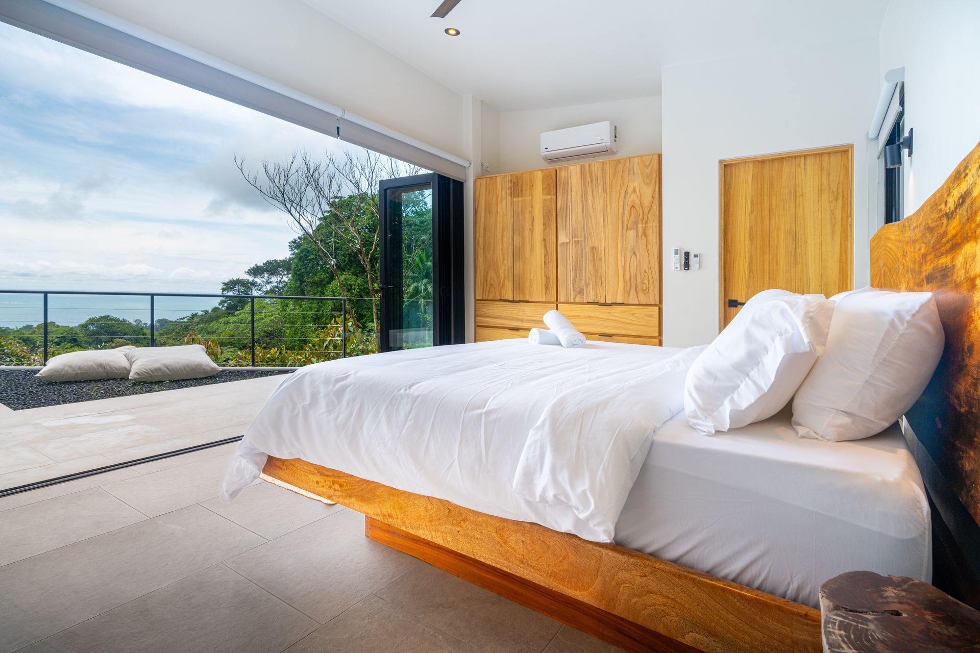 Bright modern bedroom with white bedding, wooden accents, and large open doors to a balcony and ocean view