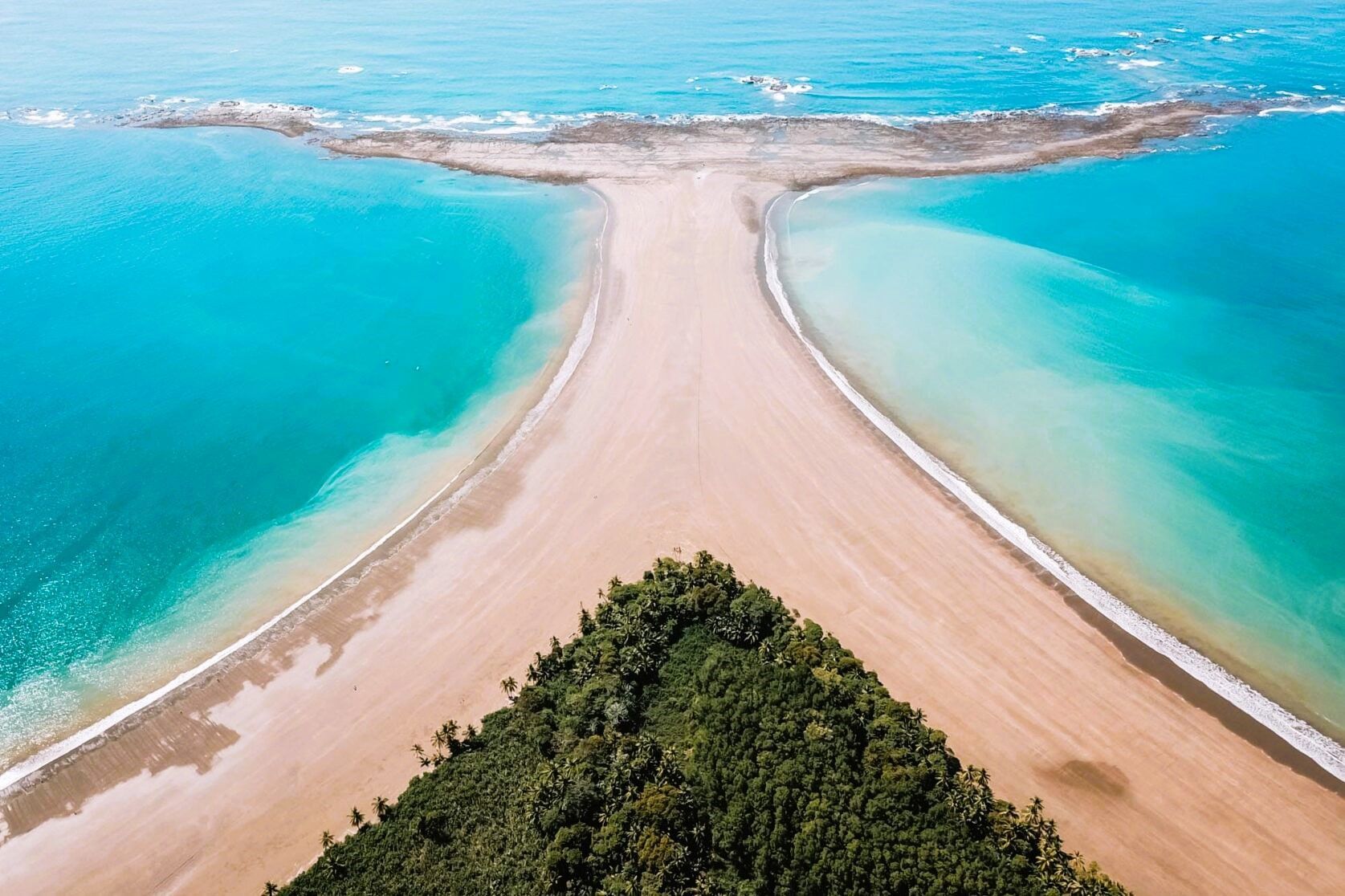 Aerial view of a sandy peninsula between turquoise waters, with a green tree line in the foreground.