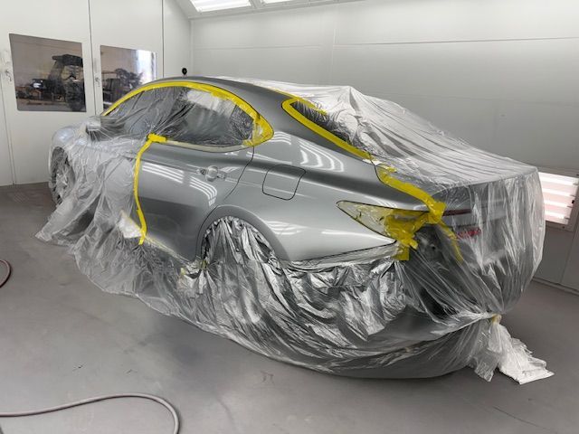 Silver car in a paint booth, partially covered with plastic sheeting and yellow tape, ready for painting.