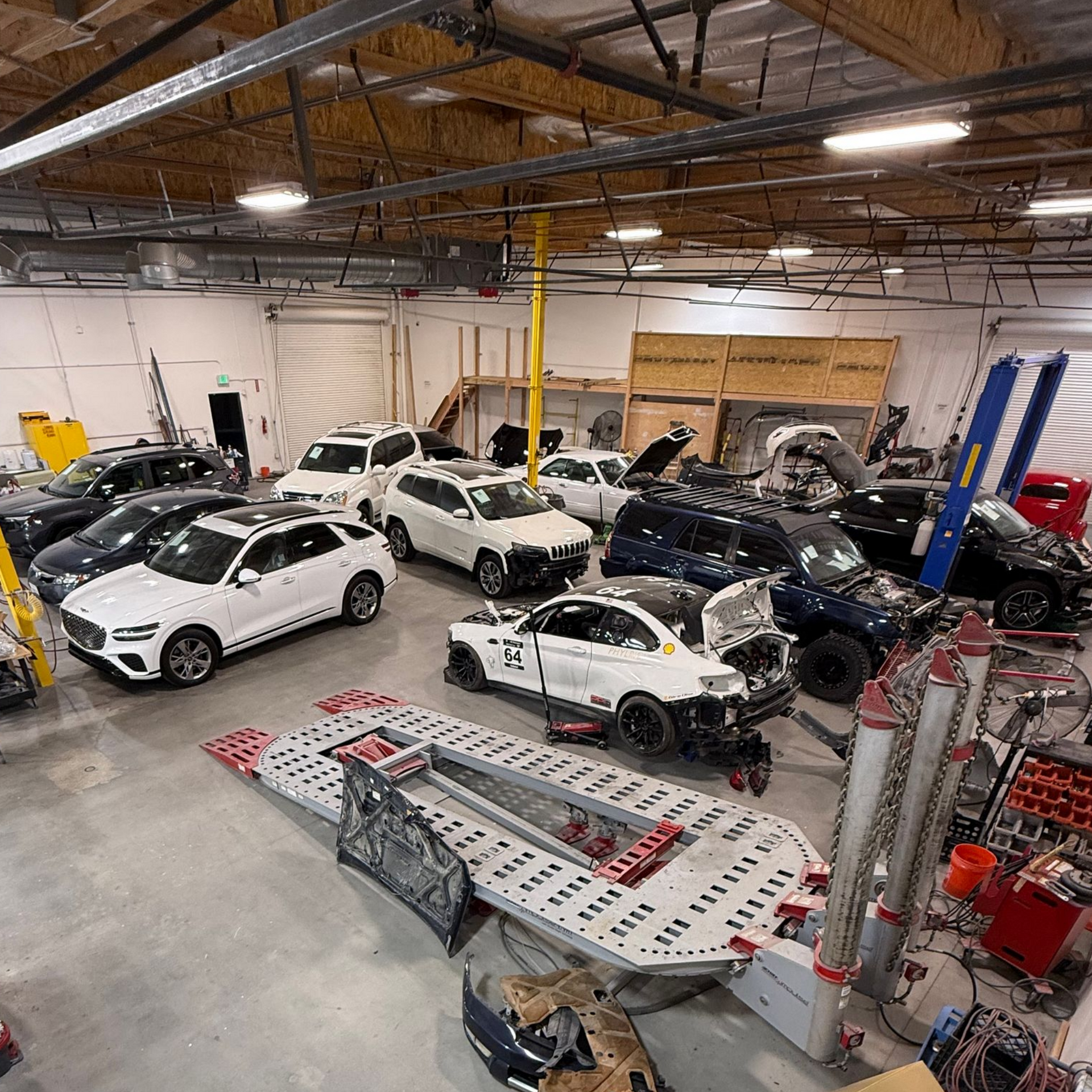 Cars in a repair shop, some damaged, around repair equipment under fluorescent lights.