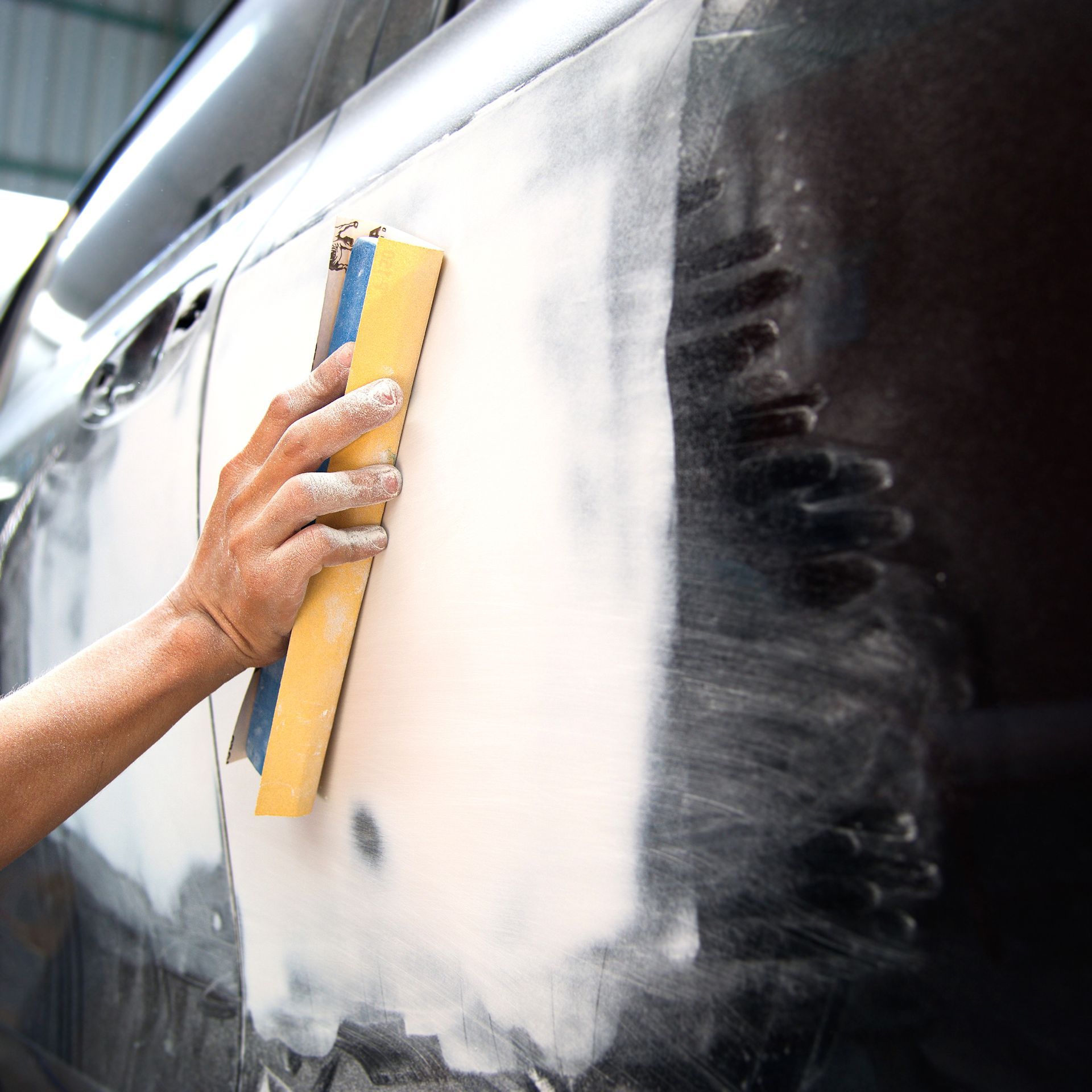 Hand sanding body filler on a black car door, smoothing the surface for paint.