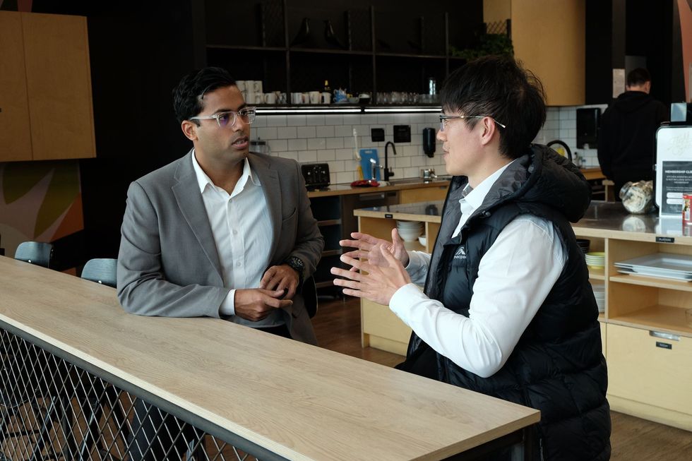 Shakti Singh and Anbin talking across a counter in a modern office kitchen, one in a gray blazer and one in a black vest.