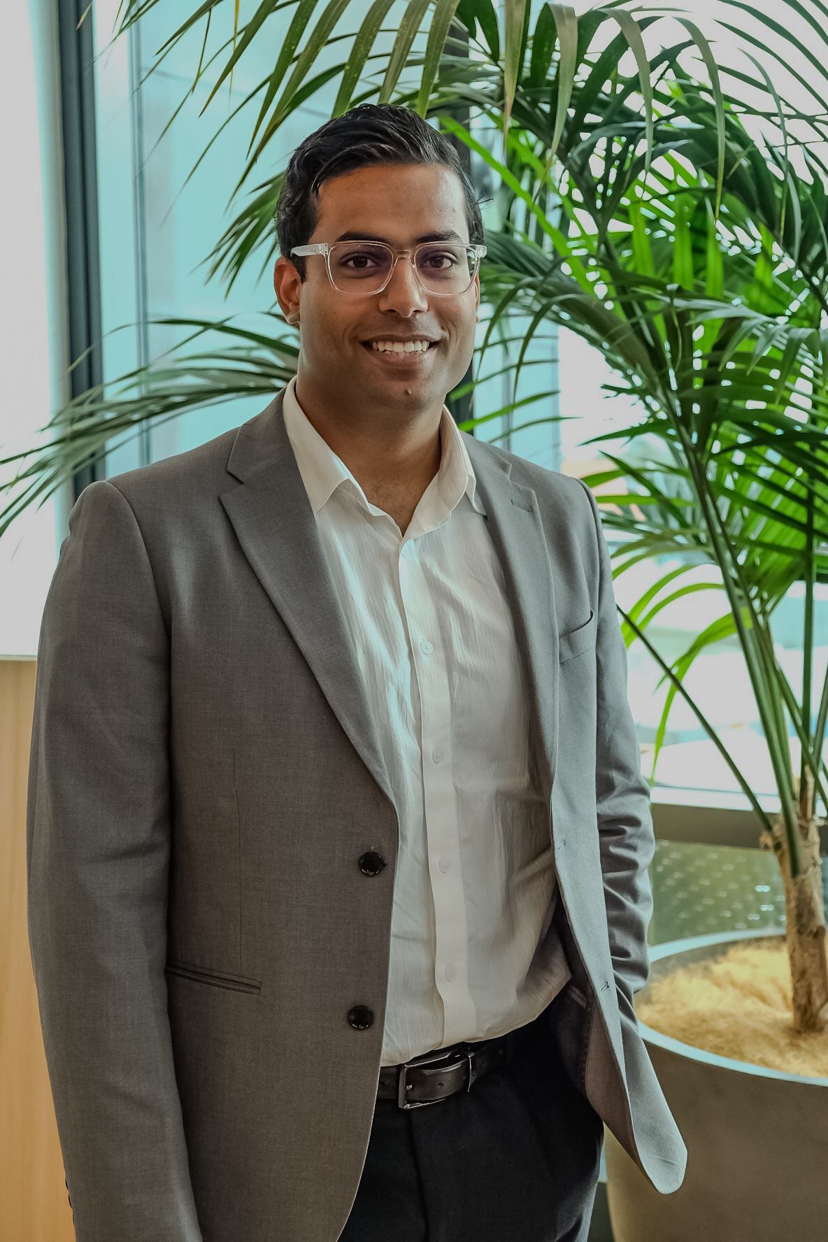 Shakti Singh in a gray blazer and white shirt standing indoors beside tall green plants, smiling at the camera.