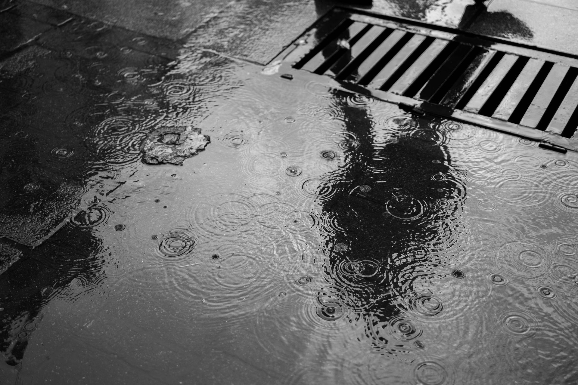 Rain puddles on a wet street beside a storm drain grate, with a person’s reflection in the water.