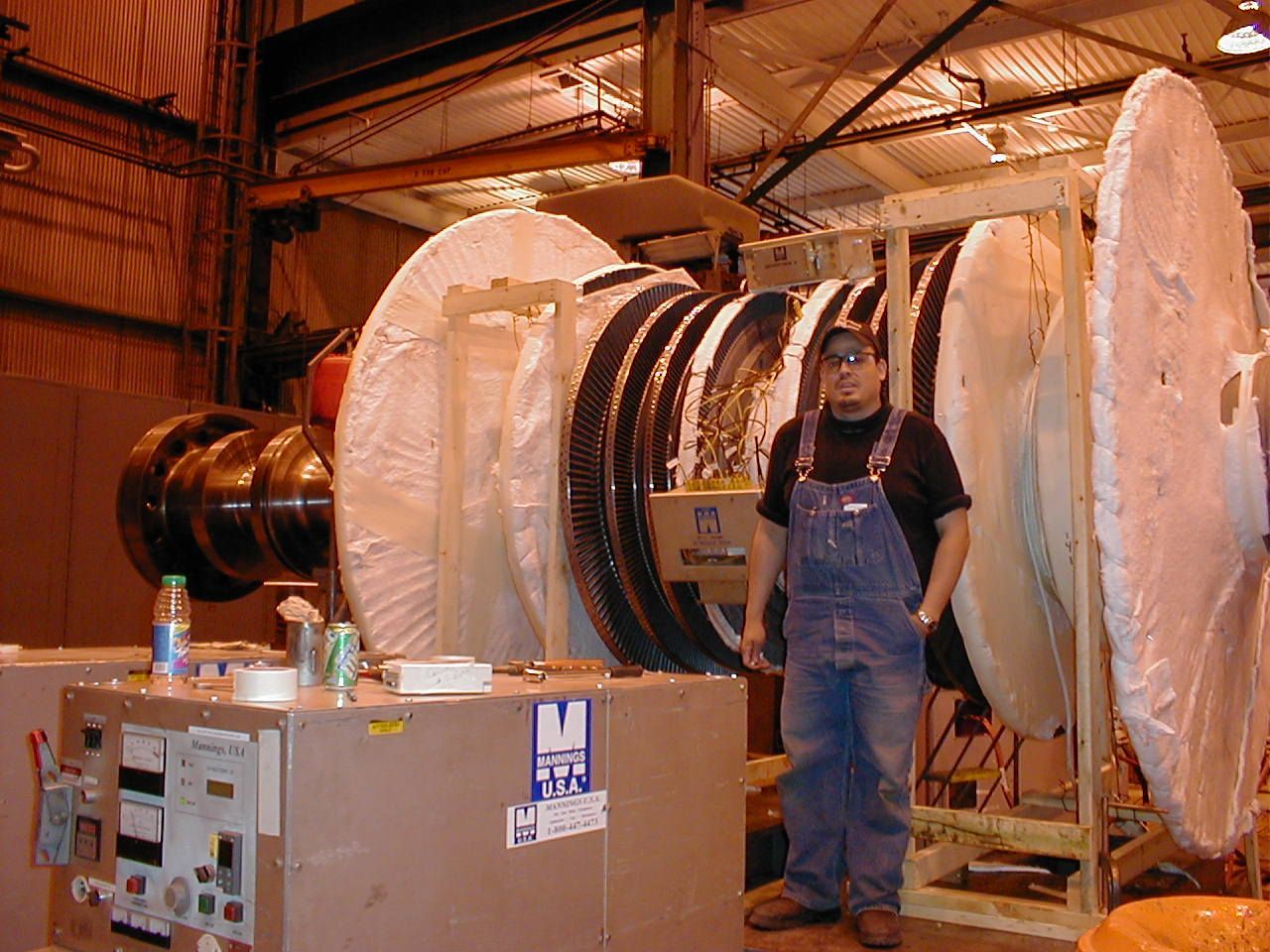 Man in overalls stands next to a large industrial turbine in a factory setting.