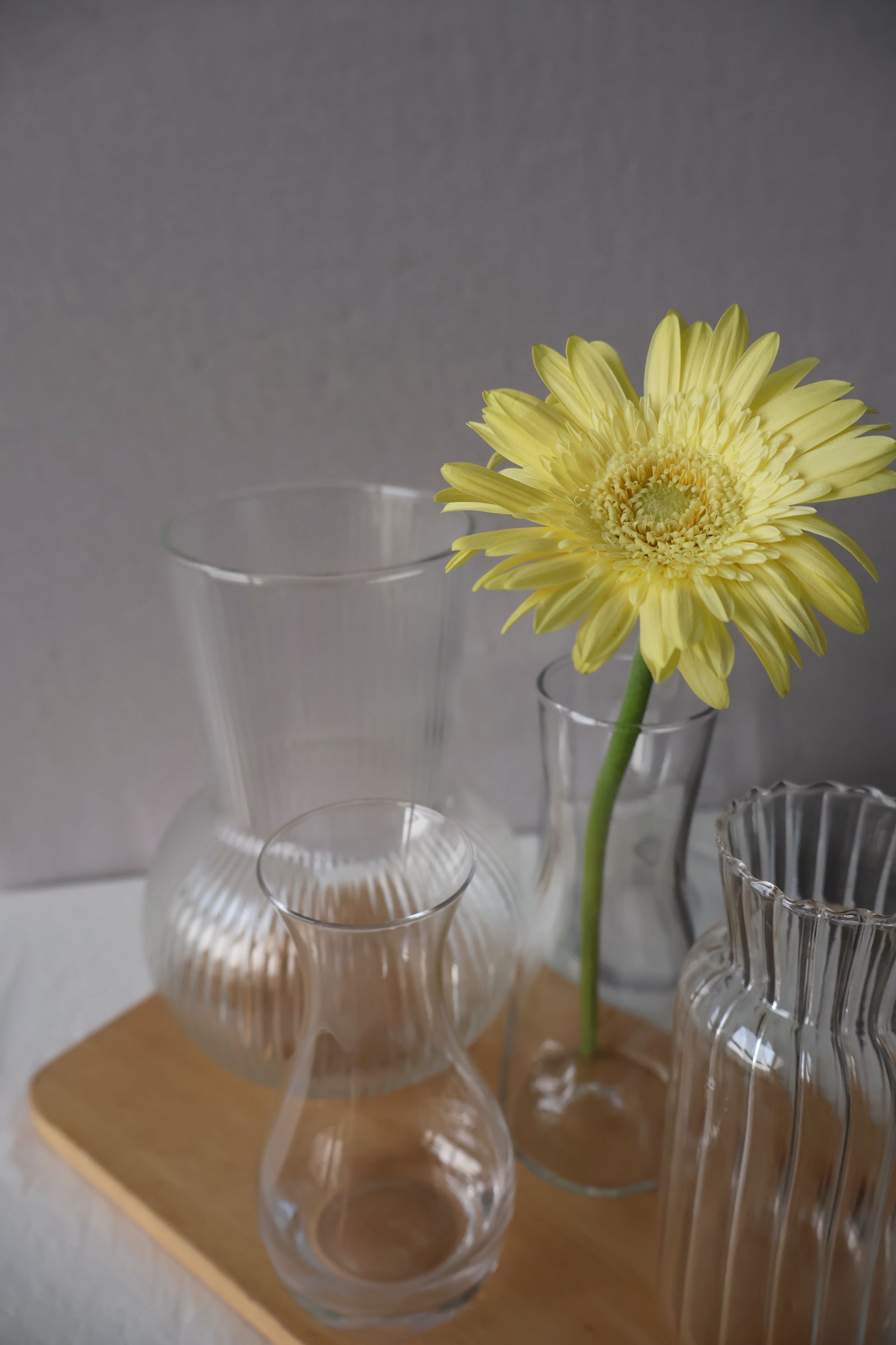 小花瓶裡插著一朵黃色的非洲菊；木托盤上放著幾個透明的棱紋玻璃花瓶。