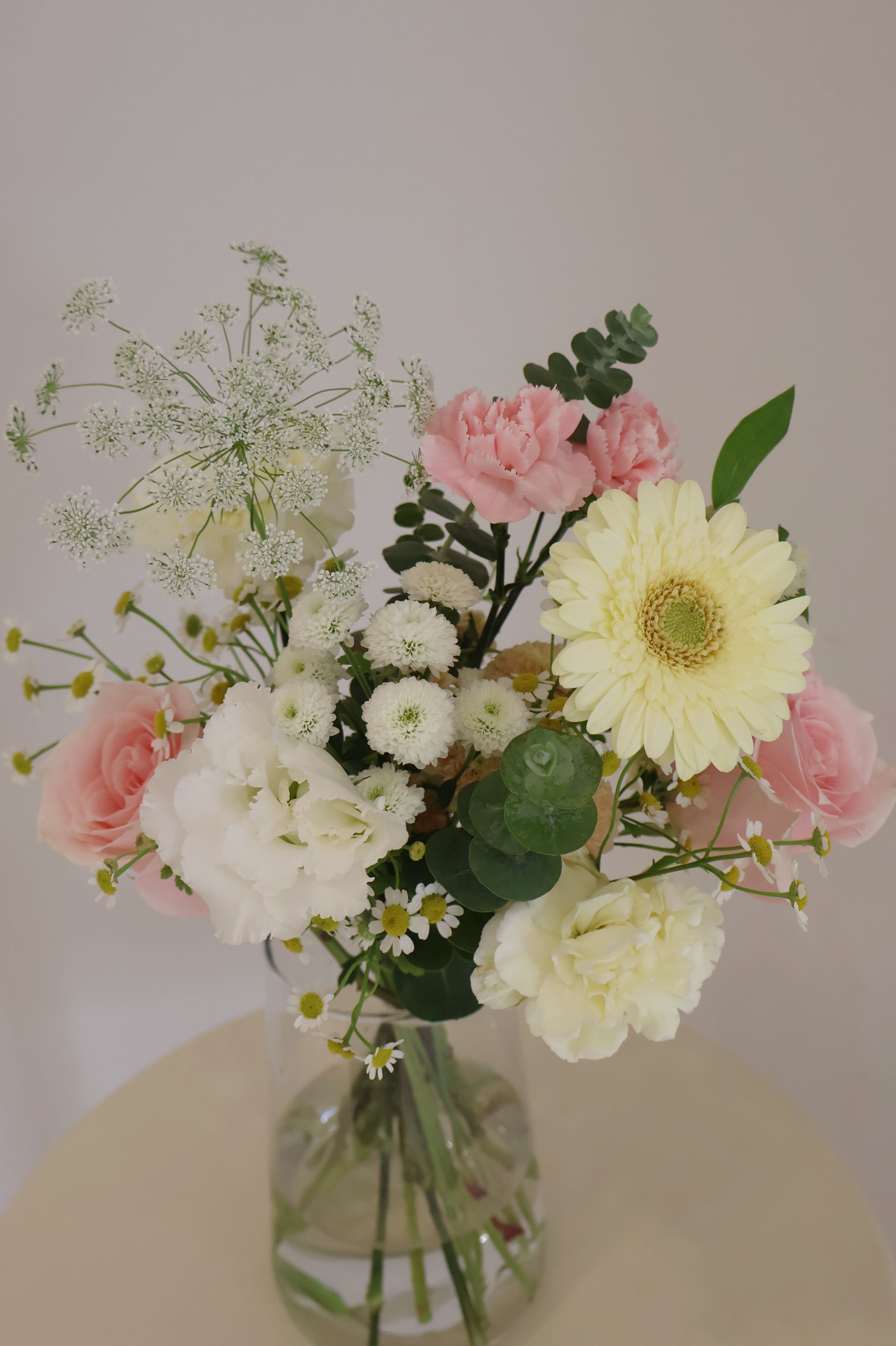 一束柔和色調的鮮花，包括非洲菊、玫瑰、康乃馨和一些配花，插在玻璃花瓶中。