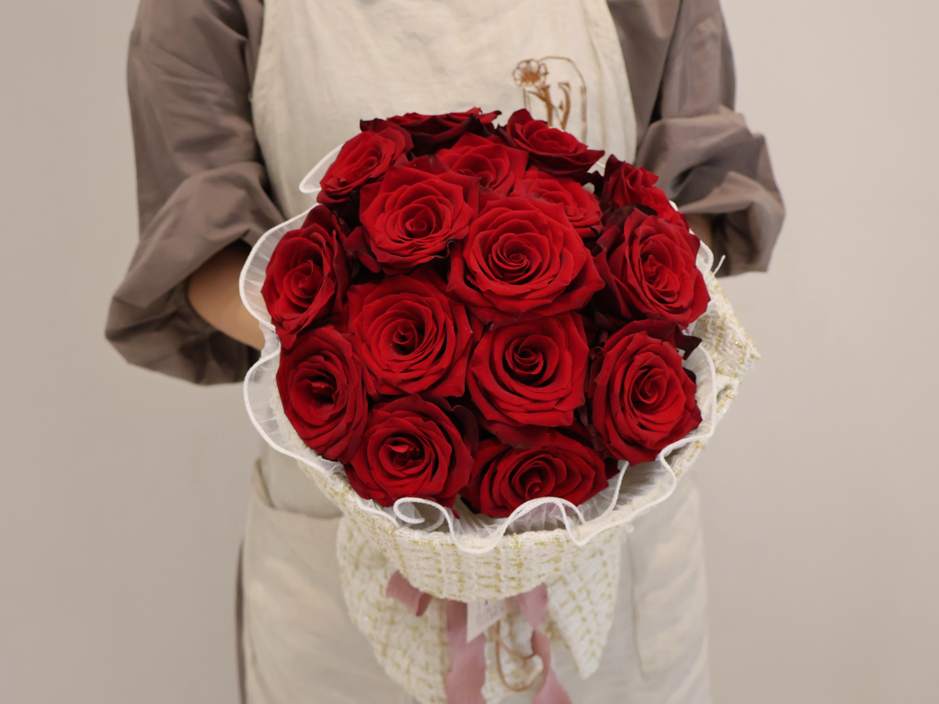 Person holding a bouquet of red roses wrapped in a lace-trimmed paper, against a plain backdrop.