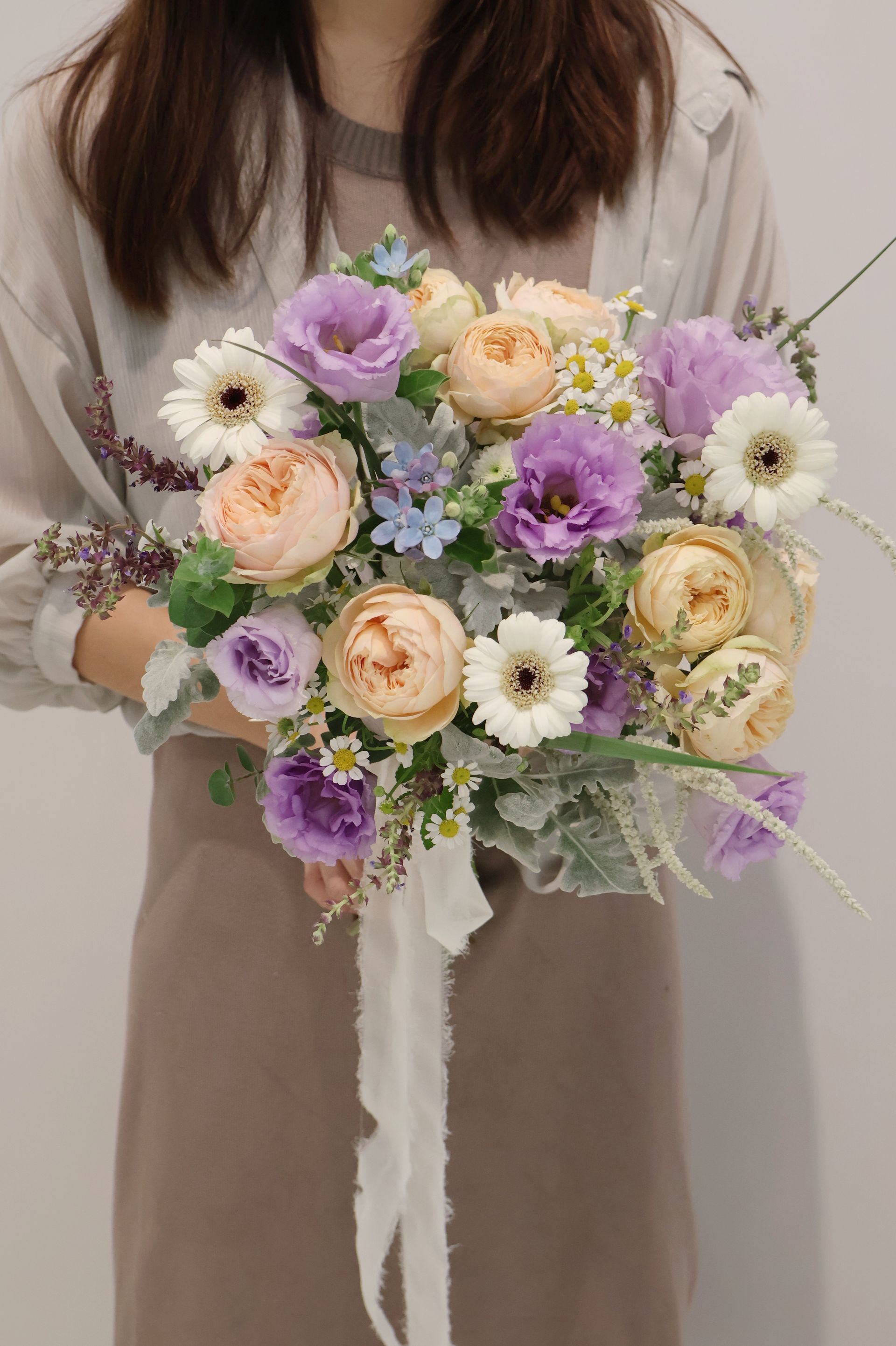 一位女士手捧一束由桃色、紫色和白色花朵組成的花束，花束上繫著白色絲帶。
