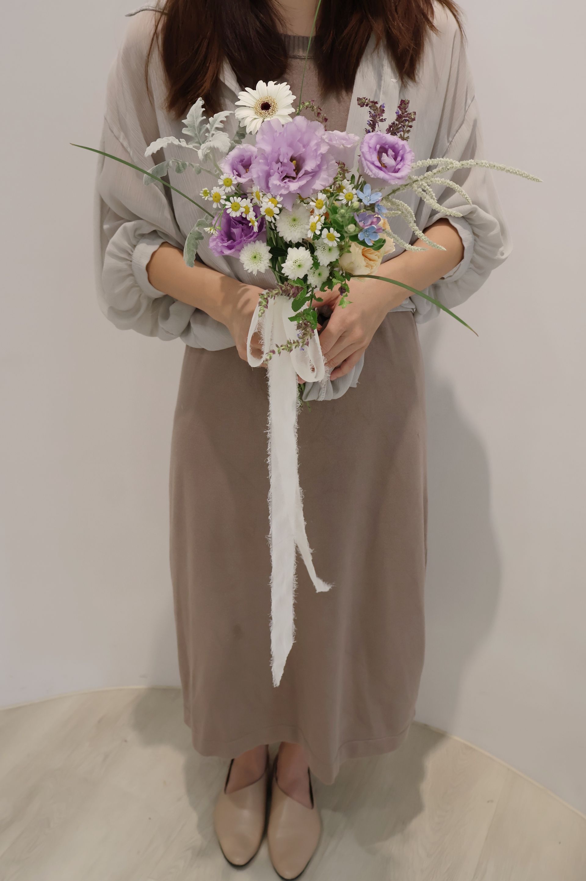 一位女子手捧一束紫白相間的鮮花，花束上繫著白色絲帶，她穿著棕色連身裙和米色襯衫。
