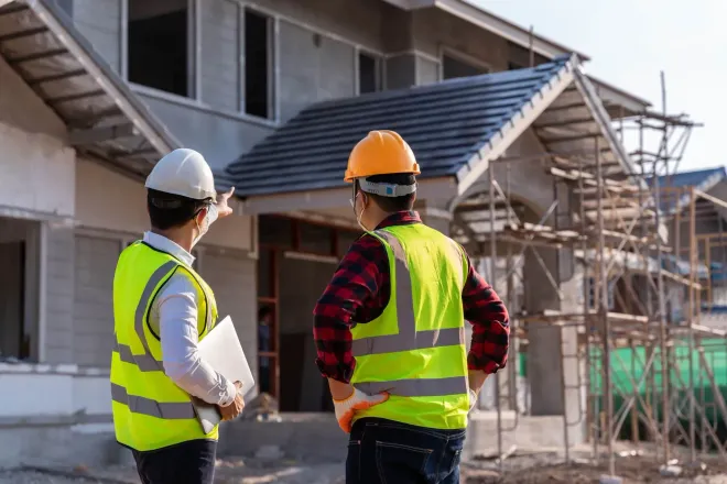 Two construction workers in high-visibility vests and hard hats stand before a house under construction.