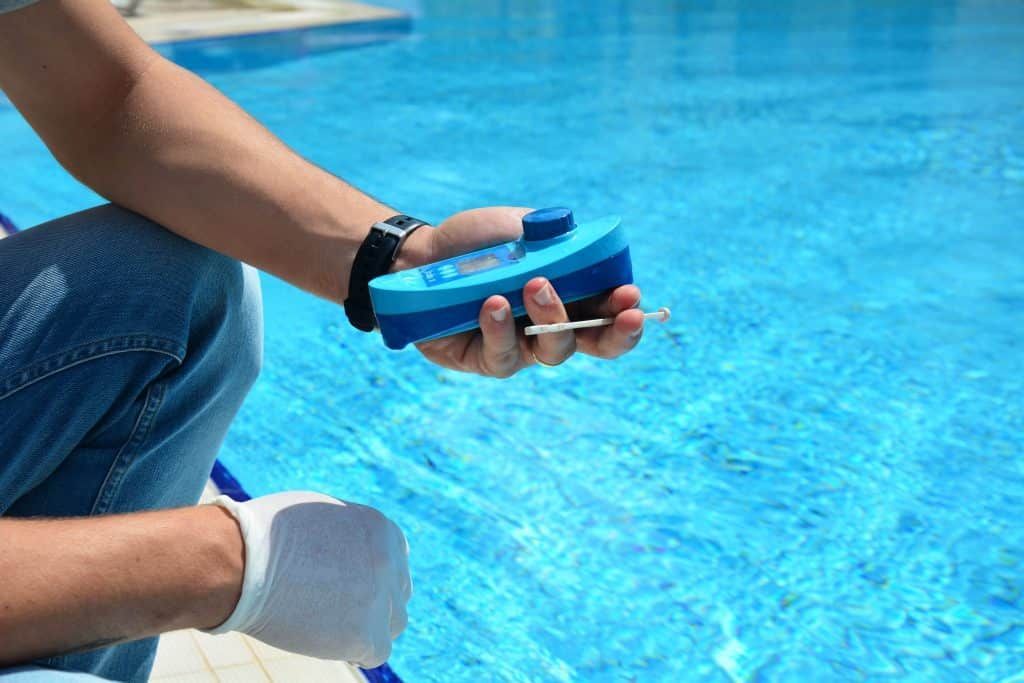 Hand holding a blue pool test kit over bright blue water, with legs by the pool edge